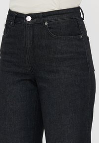 Black denim jeans with a high-waisted design, front pockets, and a silver button closure. The fabric is smooth with a subtle sheen.
