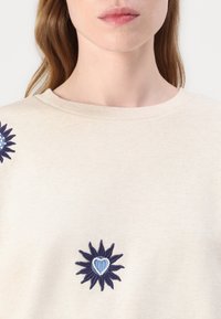 Krämfärgad sweatshirt med rund halsringning. Den har marinblå blommönsterbroderi med ett hjärta i mitten, vilket ger textur och detalj.