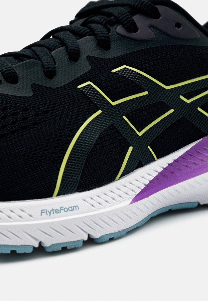 Zapatilla deportiva negra con parte superior de malla, con detalles en verde y un detalle púrpura en el talón. Plantilla blanca etiquetada "FlyteFoam."