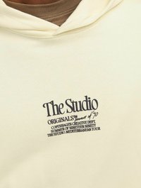 Cremefarvet hoodie med en kengurulomme. Fungerer med sort trykt tekst, der angiver mærket, oprindelse og begivenhed. Blødt stof i bomuldsblanding med glat tekstur.