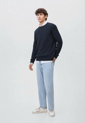 Maglione blu navy con polsini a costine, indossato sopra una camicia bianca. Jeans dritti azzurri chiari. Sneakers bianche con accenti beige. Stile minimalista.