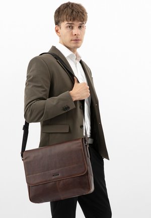 Jonge man in wit overhemd en bruin colbert met een bruine leren schoudertas tegen een effen witte achtergrond.