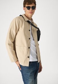 Beige corduroy jasje met lange mouwen, zwarte knopen, enkele borstzak; gedragen over een witte blouse met denim jeans. Zwarte schoudertas en zonnebril.