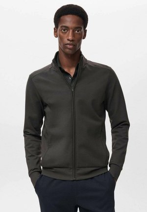 Chaqueta de color verde oscuro, con cremallera y cuello alto, hecha de un tejido texturizado. Presenta un diseño ajustado y bolsillos con cremallera ocultos.