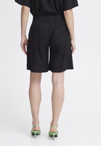 Shorts en lin noirs avec une taille élastique, arrivant aux genoux, présentant une texture lisse et une coupe simple. Associés à des sandales à talons vertes.