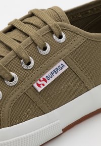 Sneaker di tela verde oliva con design a lacci, occhielli in argento, suola in gomma bianca e etichetta con logo rosso e blu sul lato.