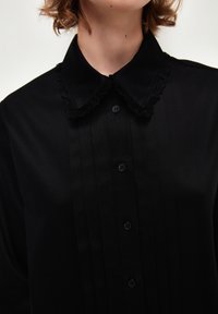 Blusa nera con davanti plissettato, colletto con pizzo e bottoni neri. Il tessuto appare liscio con una finitura opaca.
