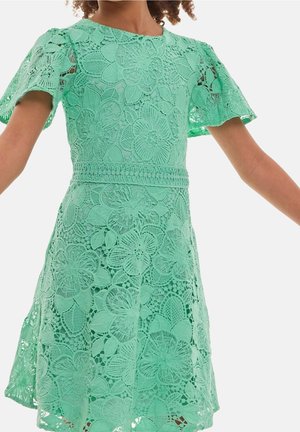 Ung person iført mintgrøn blondekjole med korte, flagrende ærmer og blomstret hæklede mønster, stående med udstrakte arme.