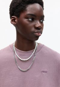 Paarse sweater met een geribbelde halslijn, met een logo. Twee kettingen: één van parels en één van zilveren kettingen, samen gelaagd.