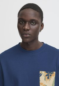 Blå bomulds t-shirt med rund hals og korte ærmer, udstyret med en lomme med blomsterpattern i gule, grønne og blå nuancer.