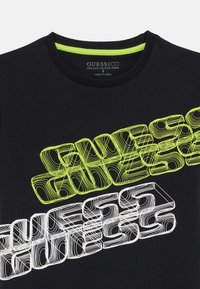 T-shirt en coton bio noir présentant un texte "GUESS" en blanc et vert fluo, en couches et en gras, avec un col rond et un tissu texturé.