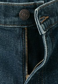 Gros plan sur un jean en denim bleu avec une fermeture éclair métallique ouverte et un bouton portant l'inscription "MNG DENIM".