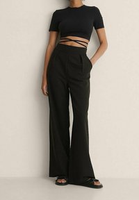 Žena oblečená v černých širokých kalhotách s vysokým pasem, černém krátkém topu s překládanými vázáními a černých sandálech, stojící vedle béžové zdi.