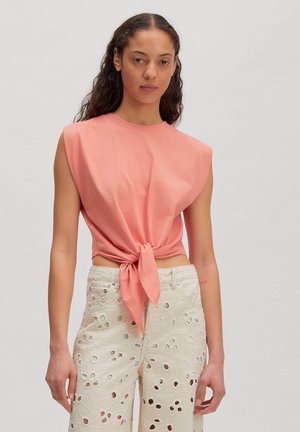 Vrouw met lang krullend haar draagt een mouwloos roze topje dat in de taille is gestrikt en een crème kleurige broek met hoge taille en bloemmotieven in de stof.