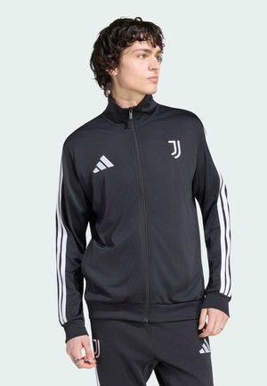 Joven con chándal negro de Adidas Juventus con rayas blancas, de pie con las manos relajadas, mirando de lado contra un fondo liso.