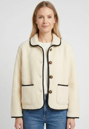 Giovane donna che indossa una giacca in pile color crema con rifiniture nere, bottoni marroni e due tasche frontali, abbinata a jeans blu e una camicia bianca.