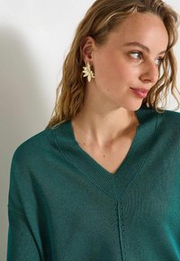 Pull vert en V avec un tricot texturé et un éclat subtil. Boucles d'oreilles florales dorées au design découpé. Le modèle a des cheveux ondulés et un maquillage neutre.