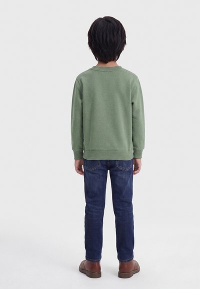 Παιδί που στέκεται με την πλάτη στραμμένη, φοράει πράσινο φούτερ, μπλε τζιν και καφέ παπούτσια σε ένα απλό φωτεινό φόντο.