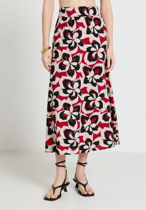 Donna che indossa una gonna midi a fiori rossa, bianca e nera, con sandali con tacco neri e cinturini e braccialetti dorati.