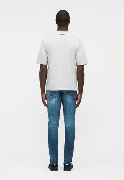 Άνδρας που στέκεται με την πλάτη προς τα εμπρός, φοράει ανοιχτό γκρι κοντομάνικο μπλουζάκι και εφαρμοστά μπλε τζιν με μαύρα παπούτσια σε λευκό φόντο.