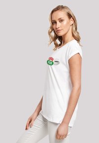 Weißes Baumwoll-T-Shirt mit hochgerollten Ärmeln, das ein farbenfrohes "Central Perk"-Logo und eine Kaffeetasse-Grafik auf der Vorderseite zeigt. Glatte Textur.