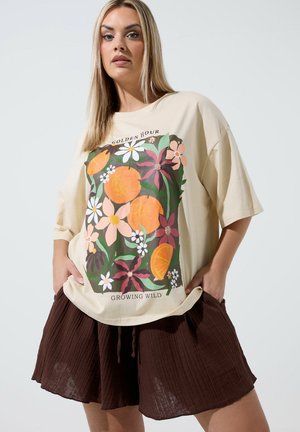 Kvinde iført beige oversized T-shirt med farverigt blomstermønster og orange tryk samt brune plisserede shorts, hænderne i lommerne, neutral baggrund.