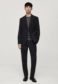 Mørkeblå habit med en tætsiddende blazer og matchende bukser, lagd over en grå sweater. Afsluttet med sorte lædersko. Minimalistisk design.