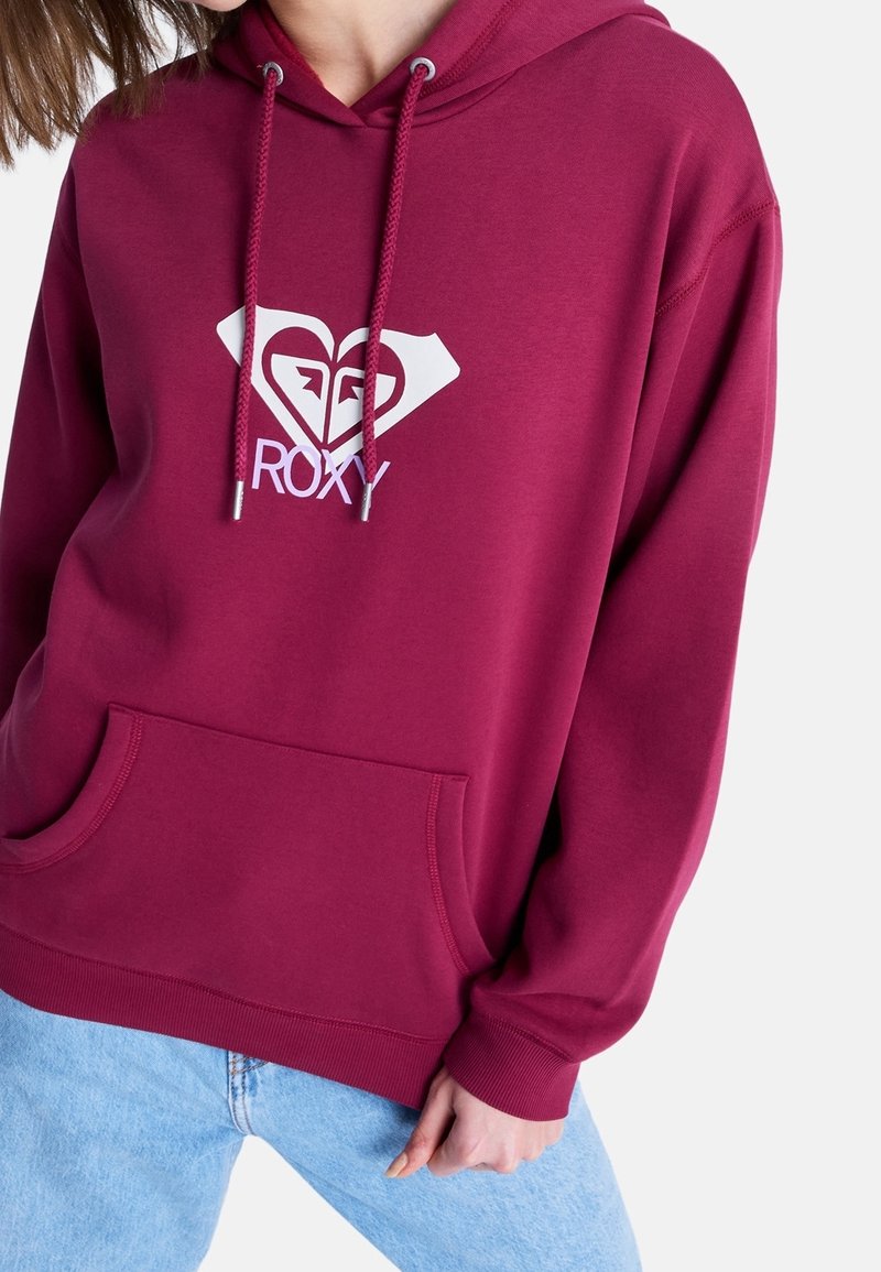 Personne portant un sweat à capuche bordeaux avec un logo Roxy blanc et un jean bleu clair sur un fond blanc uni.