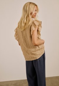 Femme blonde de profil portant un chemisier beige texturé à manches volantées et un pantalon ample foncé, sur un fond uni.
