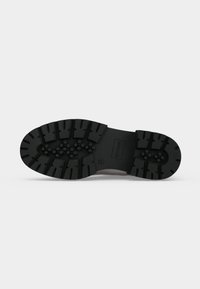 Semelle d'une botte noire avec des crampons profonds et des motifs hexagonaux et rectangulaires sur fond blanc.