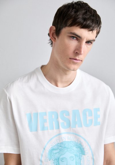 Balta kokvilnas T-krekls ar izteiksmīgu gaiši zilu tekstu "VERSACE" un centrālu grafiku ar klasisku seju un dekoratīvu apmalīti.
