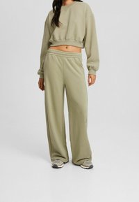 Avklippt olivgrön sweatshirt med långa ärmar; matchande vida sweatpants; mjukt tyg med elastisk midja; kombinerat med grå sneakers.