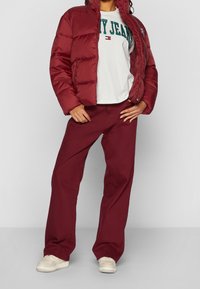 Chaqueta de plumas burdeos con un cuello alto, combinada con una camiseta gráfica blanca y pantalones de pierna ancha burdeos a juego. Zapatillas ligeras completan el look.
