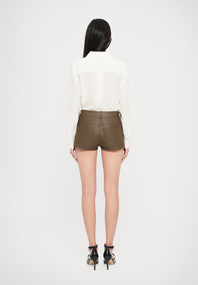 Blusa blanca transparente con un yugo en la parte posterior, combinada con shorts de cuero verde oliva. La modelo lleva sandalias de tacón negras, mostrando las piernas y los detalles del atuendo.