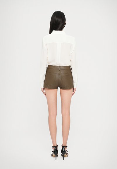 Fehér áttetsző blúz hátul yoke részlettel, olíva zöld bőrnadrággal párosítva. A modellen fekete magassarkú szandál van, kiemelve a lábakat és az öltözet részleteit.