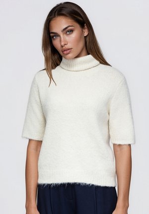 Modèle portant un pull à col roulé à manches courtes de couleur crème et un pantalon bleu marine, sur un fond uni clair.