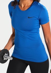 Mujer con camiseta azul de Nike levantando un dumbbell, mostrando la parte superior del cuerpo y el torso en ropa de entrenamiento contra un fondo blanco.