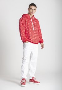 Sudadera roja con un patrón de logotipos en toda su superficie, cordones blancos, combinada con jeans blancos y zapatillas rojas con un patrón similar.