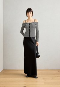 Top sans épaules rayé noir et blanc avec des boutons dorés, associé à une jupe maxi noire ajustée. Le mannequin tient un petit sac à main noir.