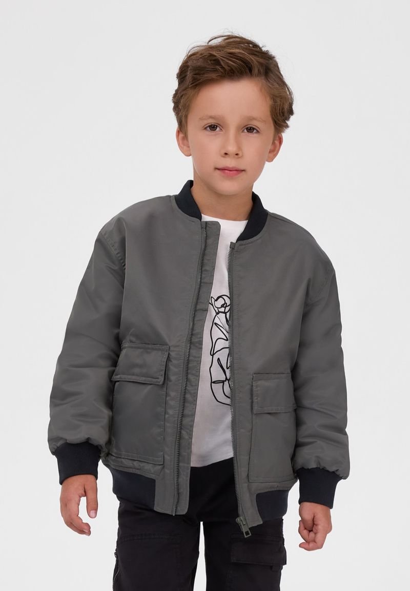 Garçon aux cheveux courts et bruns portant une veste bomber grise sur un t-shirt blanc avec un motif noir et un pantalon noir, debout devant un fond uni.