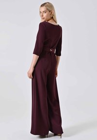 Tuta bordeaux con corpetto aderente, maniche a ¾, gambe larghe e vita con cintura caratterizzata da due bottoni dorati sul retro.