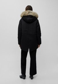 Parka invernale nera con cappuccio foderato in pelliccia, caratterizzata da un taglio lungo, tessuto testurizzato e orlo appuntito sul retro. Indossata con pantaloni neri e stivaletti.