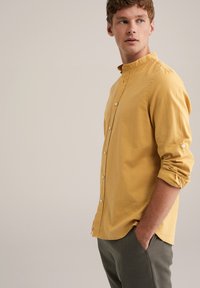Cămașă galben muștar pentru bărbați, cu guler mandarin, închidere cu nasturi și mâneci suflate, confecționată dintr-un material ușor; asortată cu pantaloni negri.