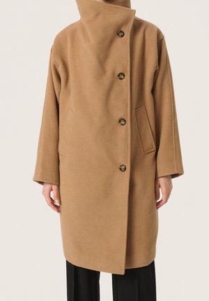Wollmantel in Kamelhaarfarbe mit großem Kragen, vier Knöpfen, Seitentaschen, getragen über schwarzen Hosen bei einer stehenden Person.