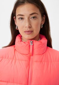 Helle rosa gepolsterte Jacke mit hohem Kragen, ausgestattet mit einem zentralen Reißverschluss und sichtbaren Nähten. Glatte Textur mit leichtem Design.