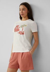 Lichtbeige t-shirt met een grafisch ontwerp van een rode en groene palmboom, ronde halslijn, korte mouwen, gecombineerd met roze sportieve shorts.