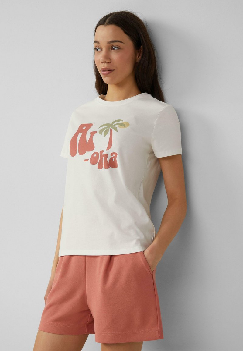 Lichtbeige t-shirt met een grafisch ontwerp van een rode en groene palmboom, ronde halslijn, korte mouwen, gecombineerd met roze sportieve shorts.
