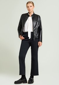 Zwarte faux leren jas met een opstaande kraag, gedragen over een witte blouse met een strik. Gecombineerd met zwarte uitlopende broek en loafers.