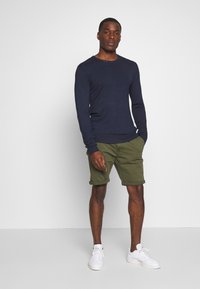 Chemise à manches longues marine assortie à un short vert olive avec des ourlets roulés. Le modèle porte des baskets blanches. Texture lisse, design simple.