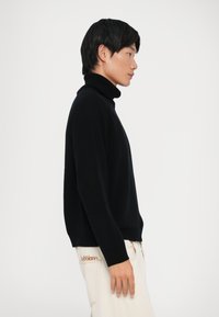 Čierny rolák s uvoľneným strihom, dlhými rukávmi a rebrovanými detailmi na golieri a manžetách, kombinovaný so svetlými nohavicami.